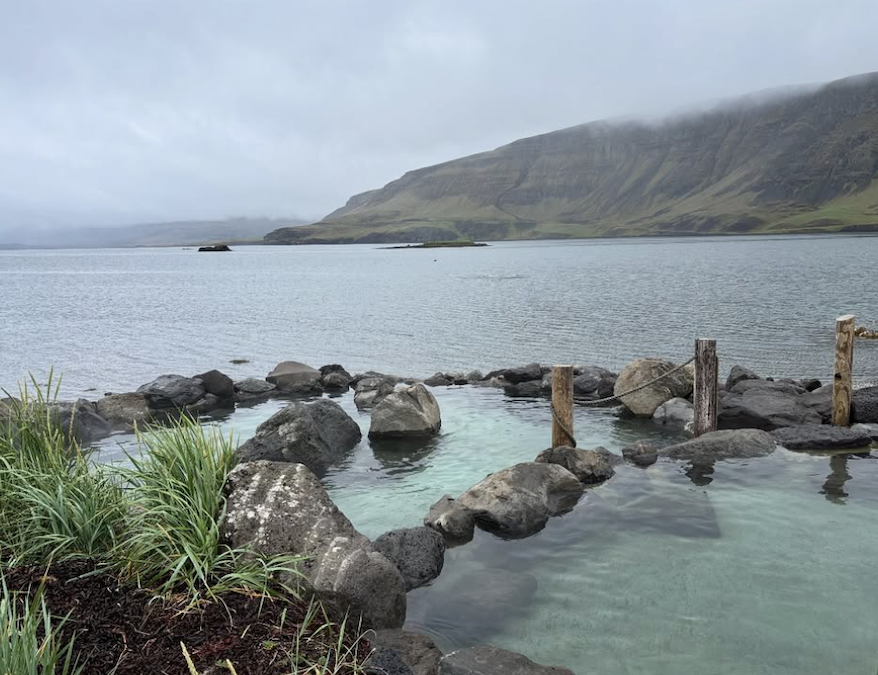 A spa in nature, with hot springs pool near the ocean, surrounded by rocks and lush greens and mountain in the distance.